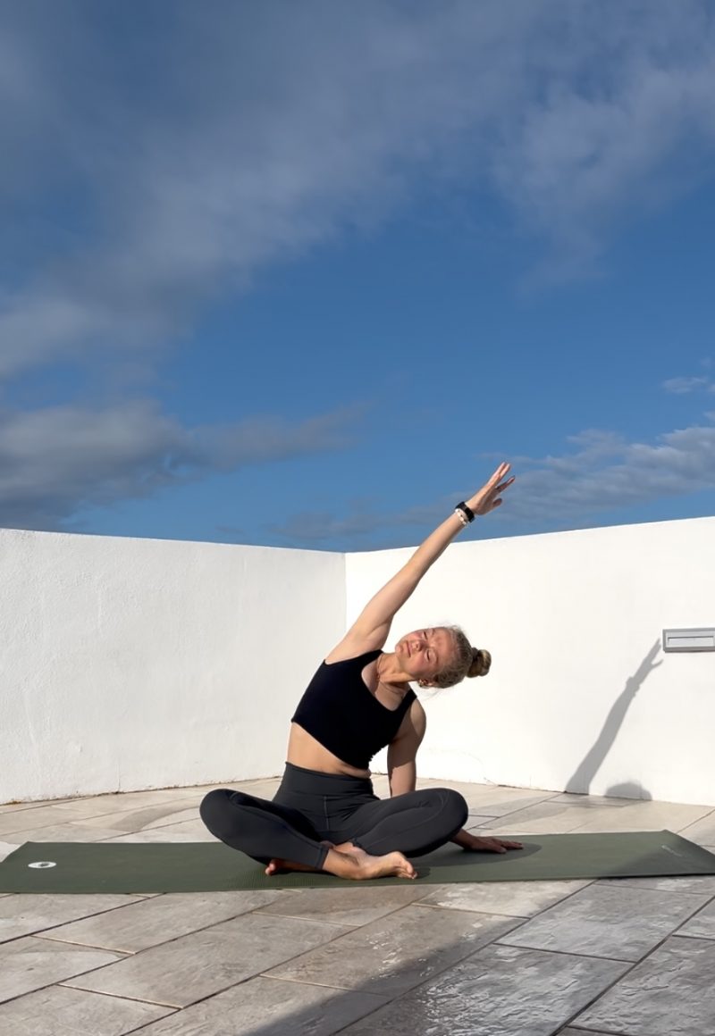 Sinah sitzend auf ihrer Yogamatte unter blauem Himmel mit leichten Sidebend